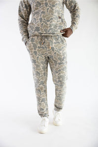 A person wearing a matching camouflage sweatshirt and jogger pants set in shades of beige, gray, and olive green, paired with white sneakers, photographed against a plain white studio background.