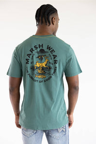 A man viewed from the back wearing a sage green t-shirt with a large yellow and black graphic print on the back that reads 'MARSH WEAR' and 'THE PURSUIT OF GOOD TIMES'.