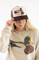 Model wearing the Mallard Duck Trucker Hat for Women in Cream and Brown, featuring a mesh back, brown adjustable snapback brim, and a detailed patch-stitch mallard duck graphic on the front.