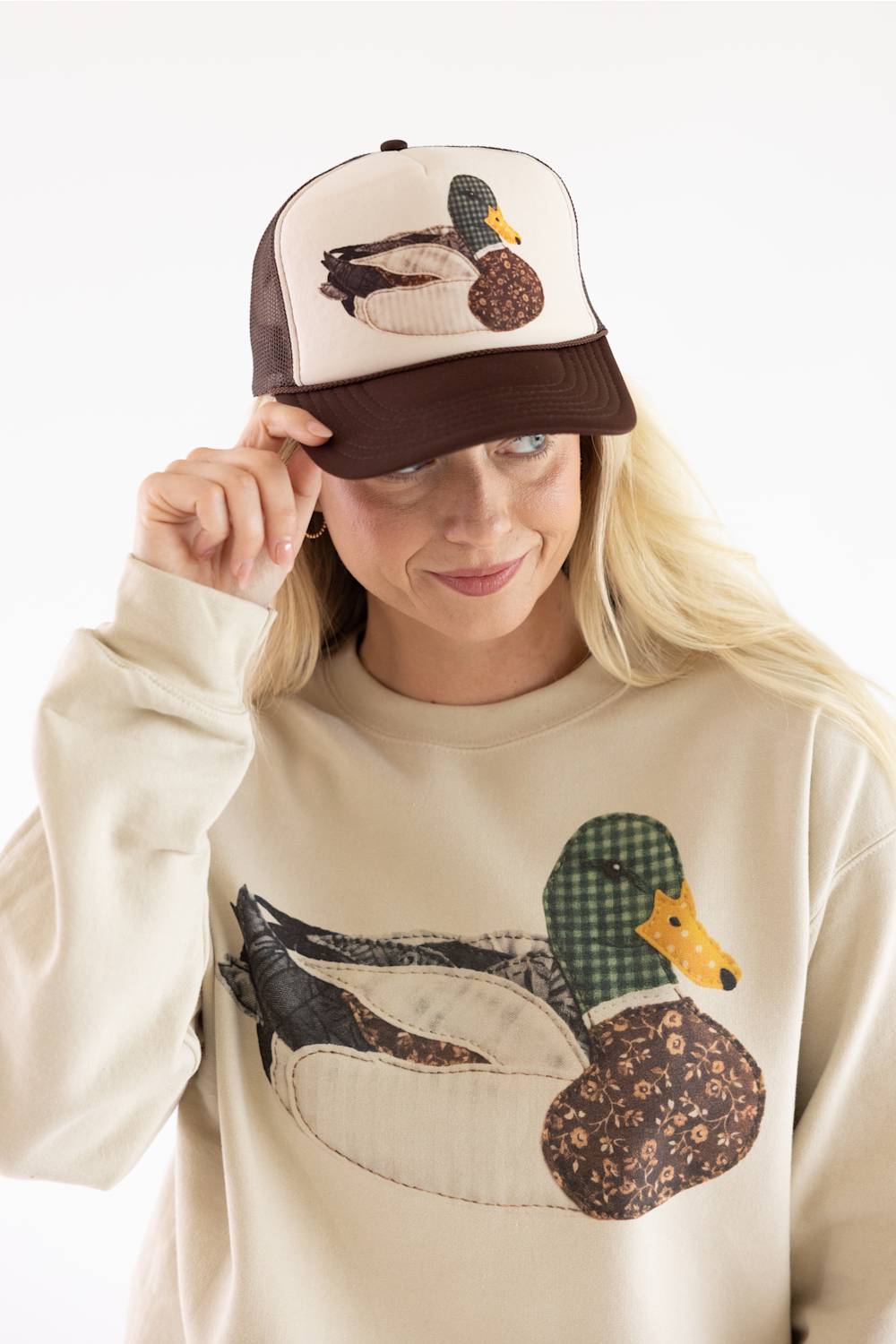 Model wearing the Mallard Duck Trucker Hat for Women in Cream and Brown, featuring a mesh back, brown adjustable snapback brim, and a detailed patch-stitch mallard duck graphic on the front.