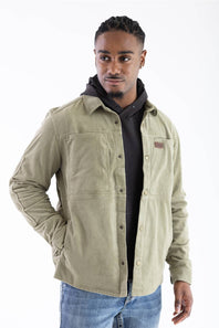 Front-facing studio shot of a model wearing the Marsh Wear Delano Cord Shacket in Sage Brush green corduroy, featuring an embossed leather brand patch on the left chest, antique brass snap closures, and dual side hand pockets, layered over a dark hoodie and styled with blue jeans.