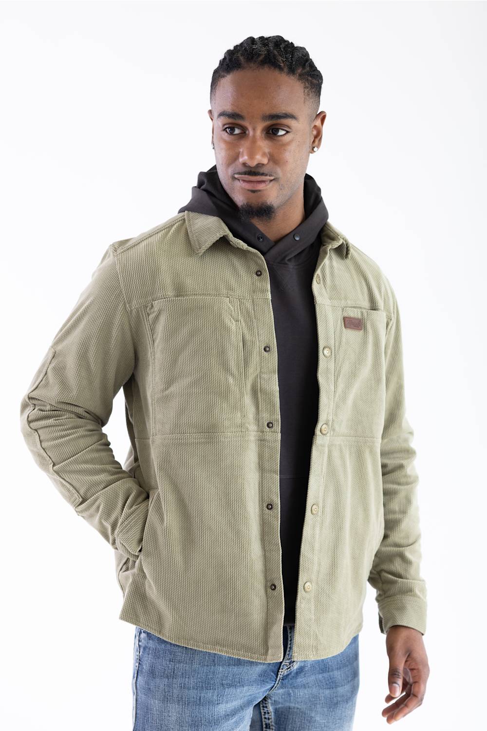 Front-facing studio shot of a model wearing the Marsh Wear Delano Cord Shacket in Sage Brush green corduroy, featuring an embossed leather brand patch on the left chest, antique brass snap closures, and dual side hand pockets, layered over a dark hoodie and styled with blue jeans.