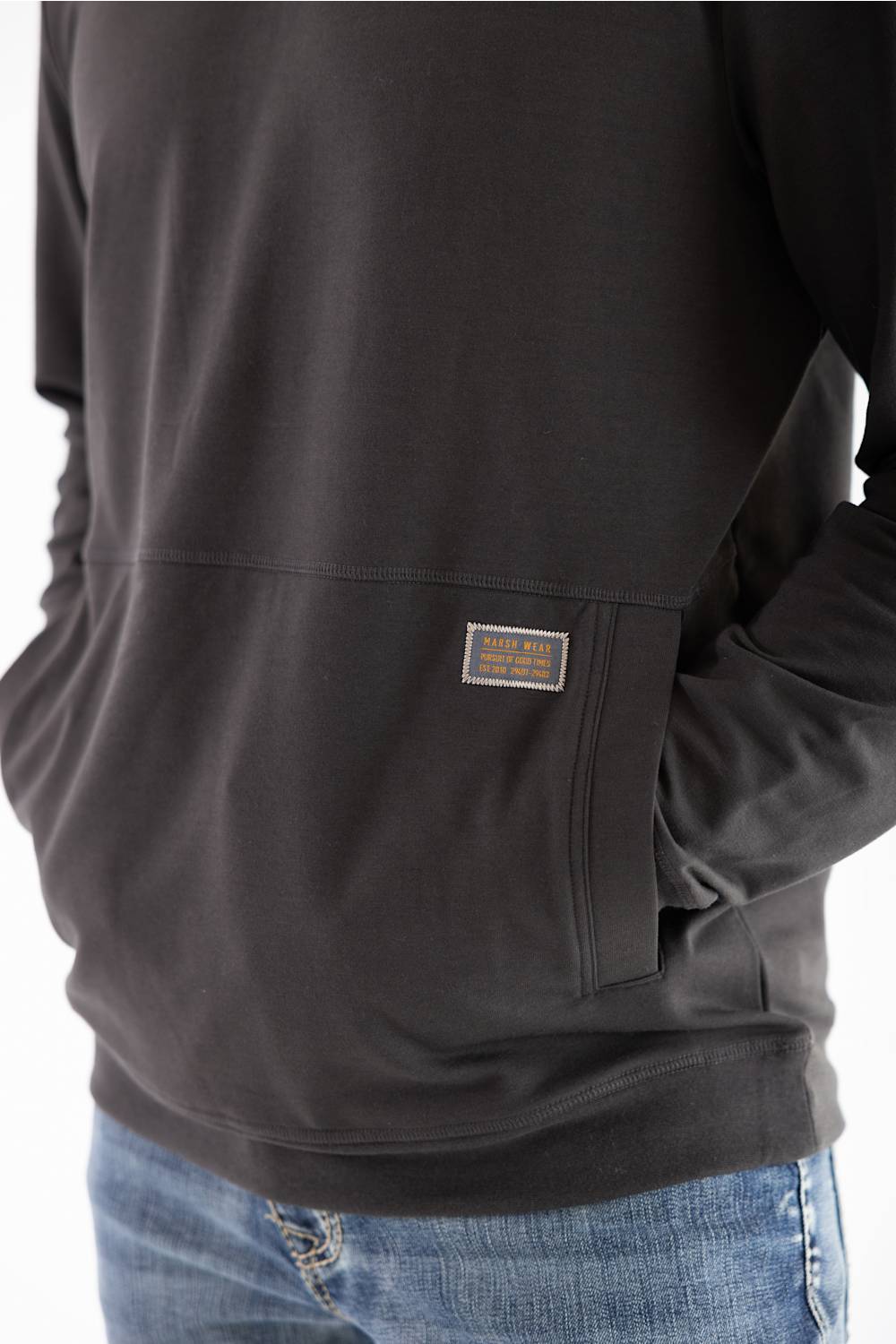 Close-up side profile of a man wearing a dark raven black long-sleeve hooded sweatshirt, highlighting the front kangaroo pocket with a small woven Marsh Wear brand patch on the left side, paired with blue jeans.