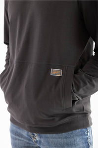 Close-up side profile of a man wearing a dark raven black long-sleeve hooded sweatshirt, highlighting the front kangaroo pocket with a small woven Marsh Wear brand patch on the left side, paired with blue jeans.