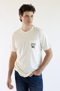 A man wearing a cream-colored short-sleeve pocket t-shirt with a black and brown 'MARSH' graphic on the chest, paired with dark blue jeans, against a plain white background.
