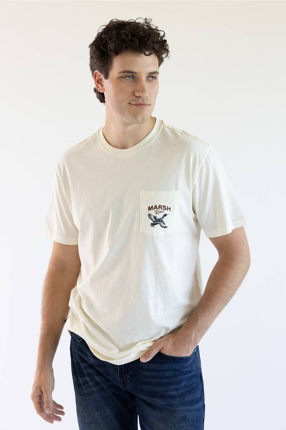 A man wearing a cream-colored short-sleeve pocket t-shirt with a black and brown 'MARSH' graphic on the chest, paired with dark blue jeans, against a plain white background.