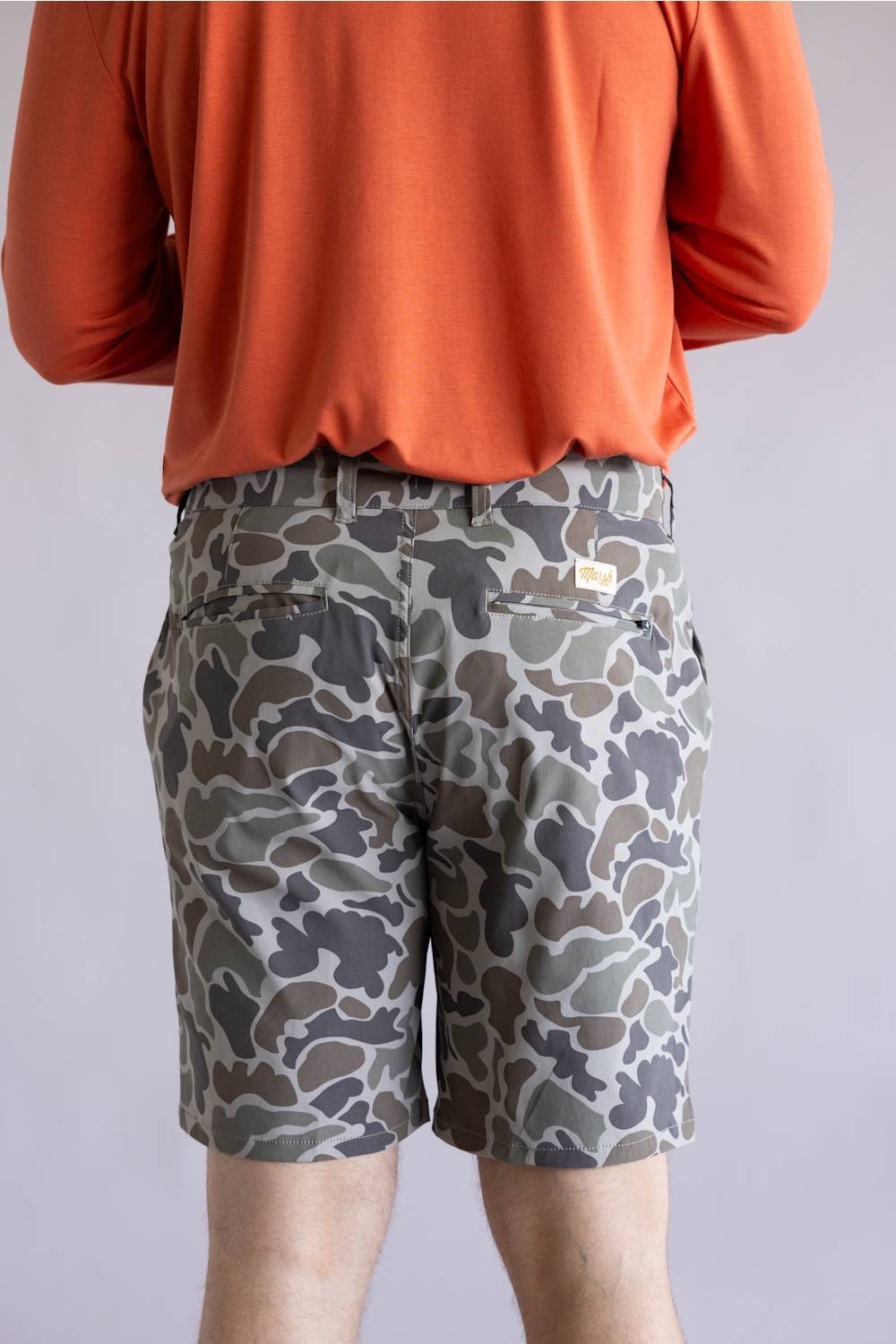 Marsh Wear Prime Hagood 8” Shorts for Men in Green Mallard Camo; Person wearing camouflage shorts and an orange long-sleeve shirt on a light gray background