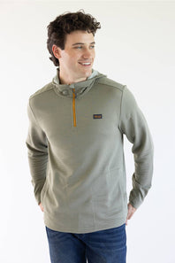 A man wearing a Marshwear hooded quarter-zip pullover in a textured sage green fabric with a small orange zipper and a small Marshwear  logo patch on the chest, paired with blue jeans against a plain white background.