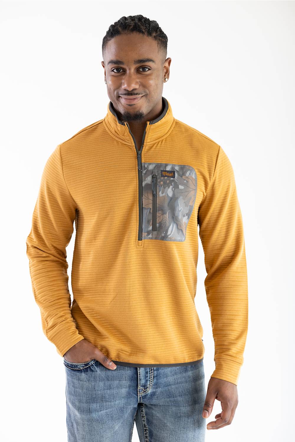Front portrait studio shot of a model smiling while wearing the Marsh Wear Sullivan Quarter Zip Pullover in Buckthorn Brown, featuring a textured ribbed design, dark grey inner stand-up collar, front quarter-zip closure, and a leaf-patterned camouflage chest zip pocket with an orange "Marsh Wear" logo patch, styled with blue jeans.