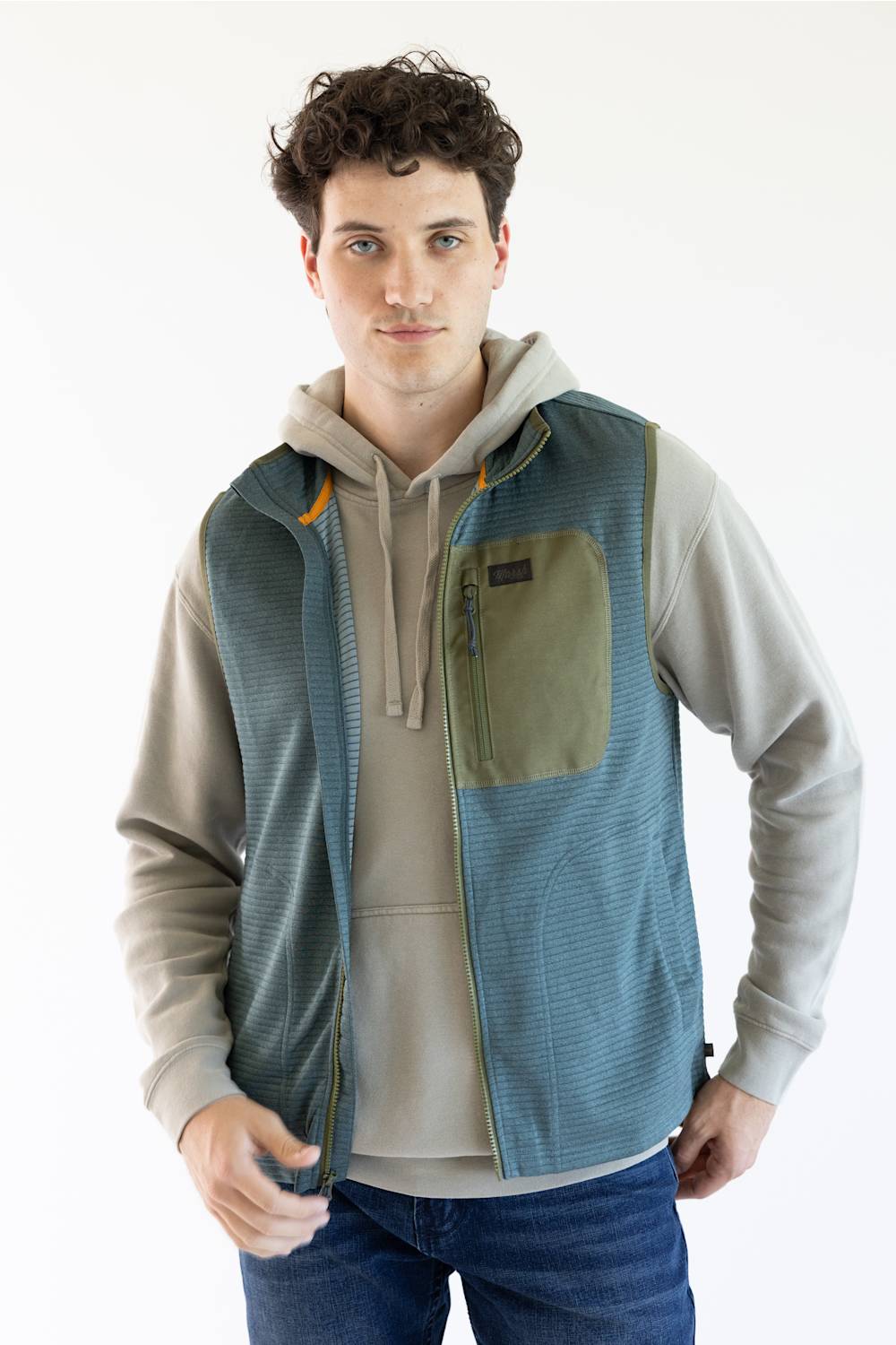 A man wearing a blue-green textured zip-up vest over a beige hoodie and blue jeans, photographed against a clean white studio background.