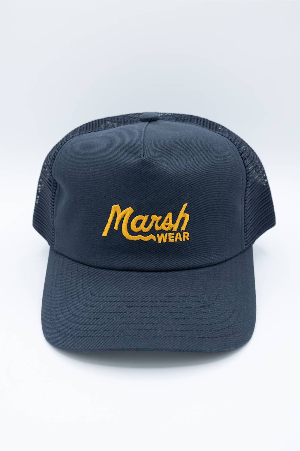 A man wearing a dark navy blue trucker hat with yellow embroidered 'Marsh Wear' text, paired with a light beige hoodie, against a plain white background.