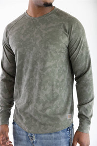 Front three-quarter close-up view of a model wearing the long-sleeve Marsh Wear Surfside Waffle Knit Shirt in Fossil Foxhole Camo green, showcasing the textured waffle-knit fabric, crew neck, raglan sleeves, and a small woven logo patch near the hem, styled with blue jeans.