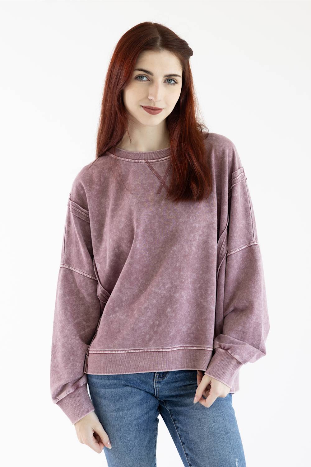 A woman with red hair wearing a faded dusty rose/mauve crewneck sweatshirt and blue jeans, standing against a plain white studio background.