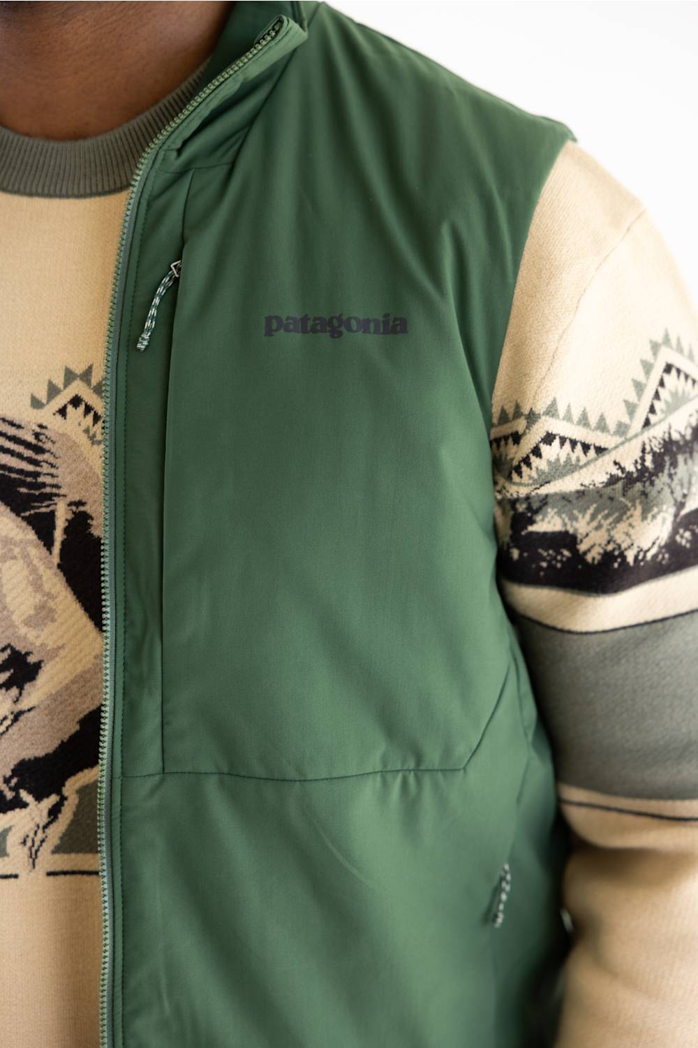 Close-up of a person wearing a Patagonia green zip-front vest over a beige and gray patterned sweater, with the Patagonia logo visible on the chest.