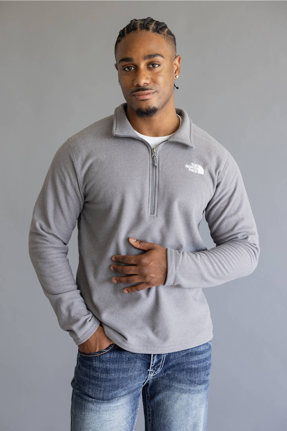 A man wearing a light gray quarter-zip fleece pullover by The North Face, paired with blue jeans, standing against a solid gray studio background.