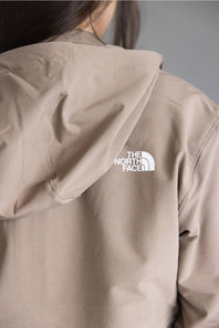 Close-up back view of a person wearing a light beige hooded jacket with a white The North Face logo on the upper back, against a plain gray background.