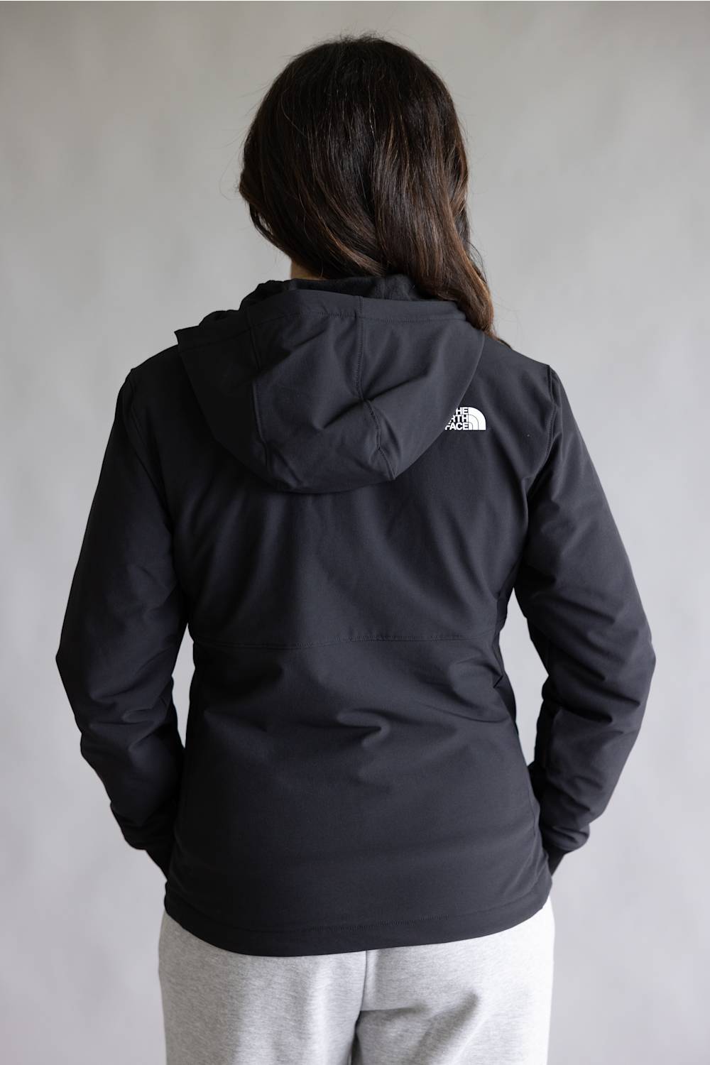 A person viewed from the back wearing a black hooded jacket with a small white The North Face logo on the upper back, paired with light gray sweatpants against a plain light gray background.