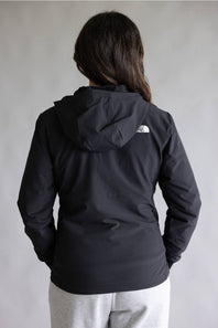 A person viewed from the back wearing a black hooded jacket with a small white The North Face logo on the upper back, paired with light gray sweatpants against a plain light gray background.