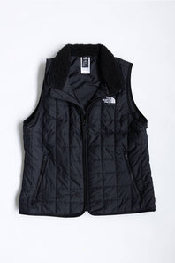 A black quilted puffer vest with a black sherpa collar, featuring a front zipper, zippered side pockets, and a white embroidered logo of The North Face on the left chest, displayed against a plain white background.