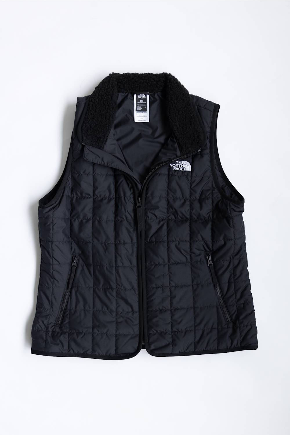 A black quilted puffer vest with a black sherpa collar, featuring a front zipper, zippered side pockets, and a white embroidered logo of The North Face on the left chest, displayed against a plain white background.