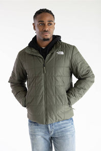 A man wearing an olive green puffer jacket by The North Face, styled with a black hoodie and blue jeans, photographed against a plain white studio background.