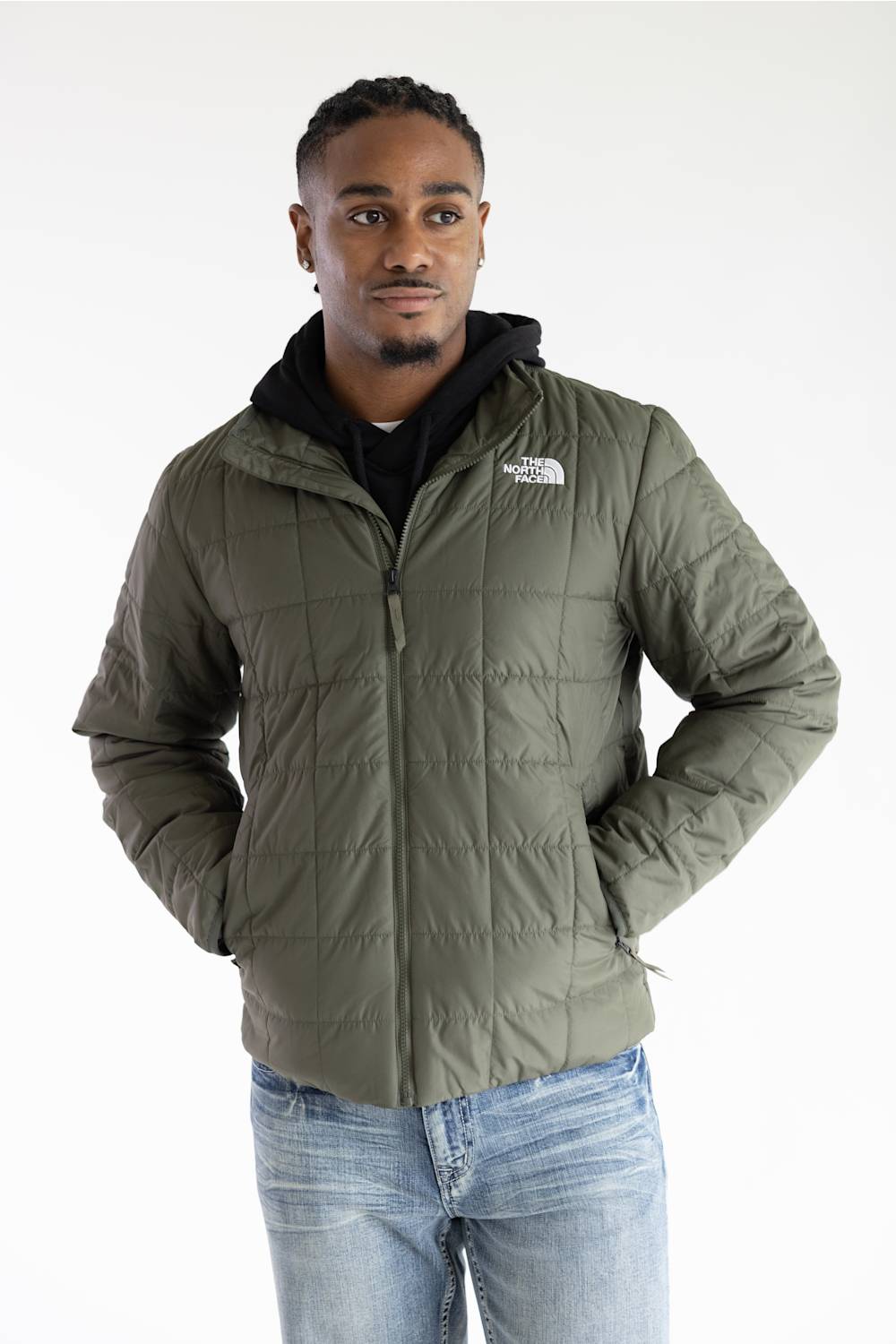 A man wearing an olive green puffer jacket by The North Face, styled with a black hoodie and blue jeans, photographed against a plain white studio background.