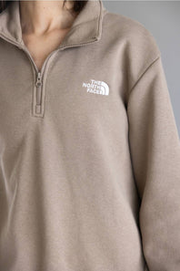 Close-up of a person wearing a taupe quarter-zip pullover sweatshirt with a white embroidered The North Face logo on the left chest.