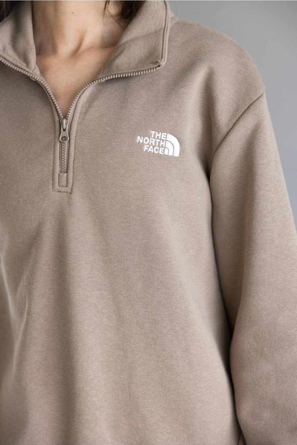 Close-up of a person wearing a taupe quarter-zip pullover sweatshirt with a white embroidered The North Face logo on the left chest.