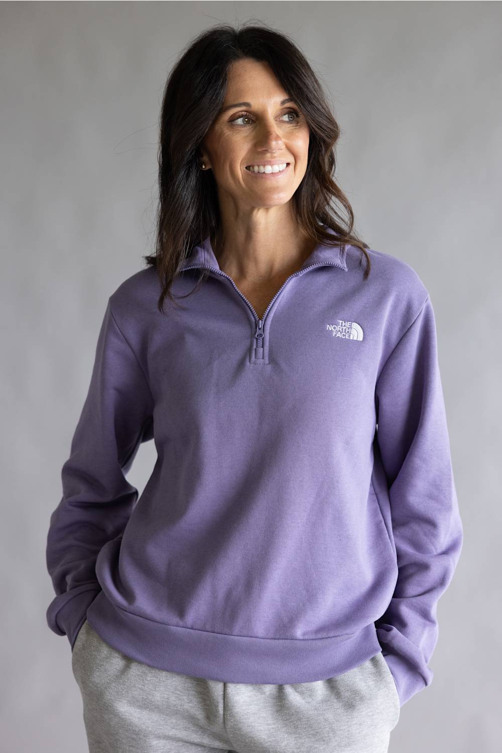 A woman wearing a purple quarter-zip sweatshirt by The North Face, paired with light gray sweatpants, standing against a neutral gray studio background.