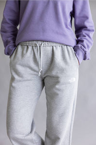 A person wearing light heather gray sweatpants with a white drawstring and a small white The North Face logo on the left thigh, paired with a lavender sweatshirt against a plain light gray background.