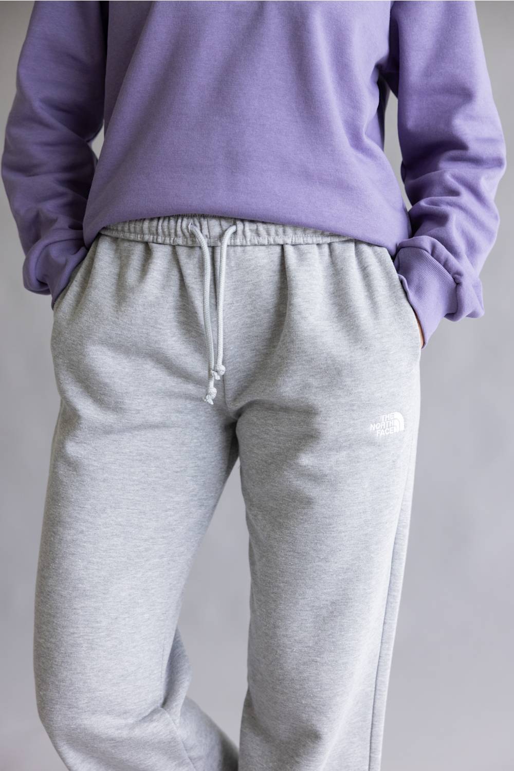 A person wearing light heather gray sweatpants with a white drawstring and a small white The North Face logo on the left thigh, paired with a lavender sweatshirt against a plain light gray background.
