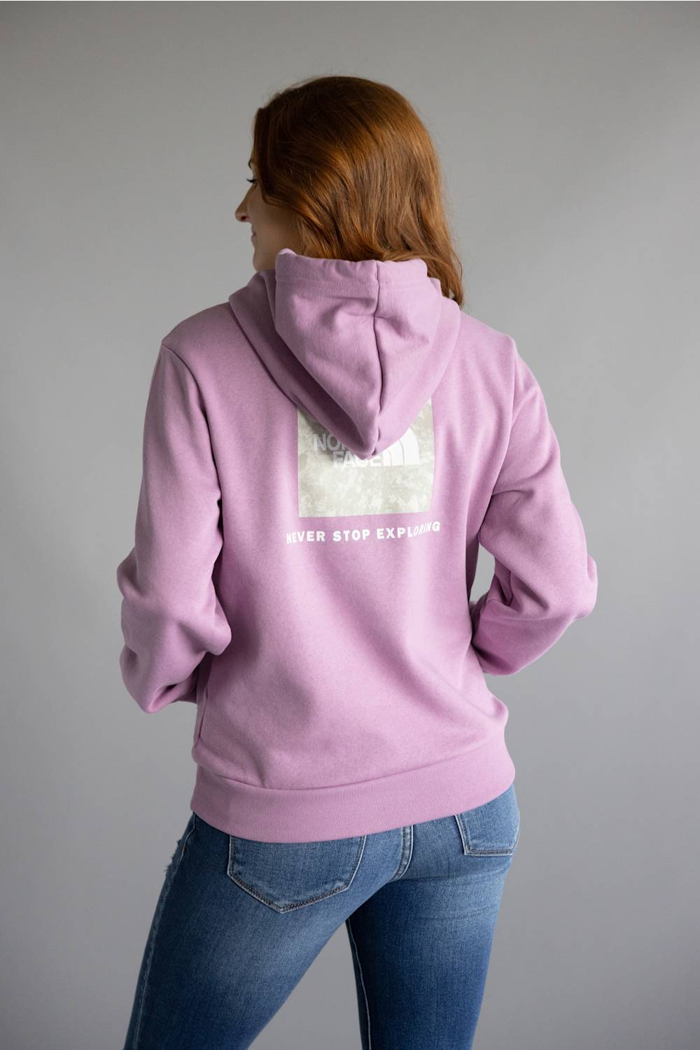 A woman with red hair is shown from the back wearing a lavender purple hoodie with a white graphic print on the back, paired with blue jeans, against a solid gray studio background.