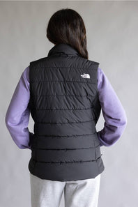 A person shown from the back wearing a black quilted puffer vest over a lavender long-sleeve shirt and light gray sweatpants, against a plain light gray studio background. The The North Face logo is visible on the upper back of the vest.