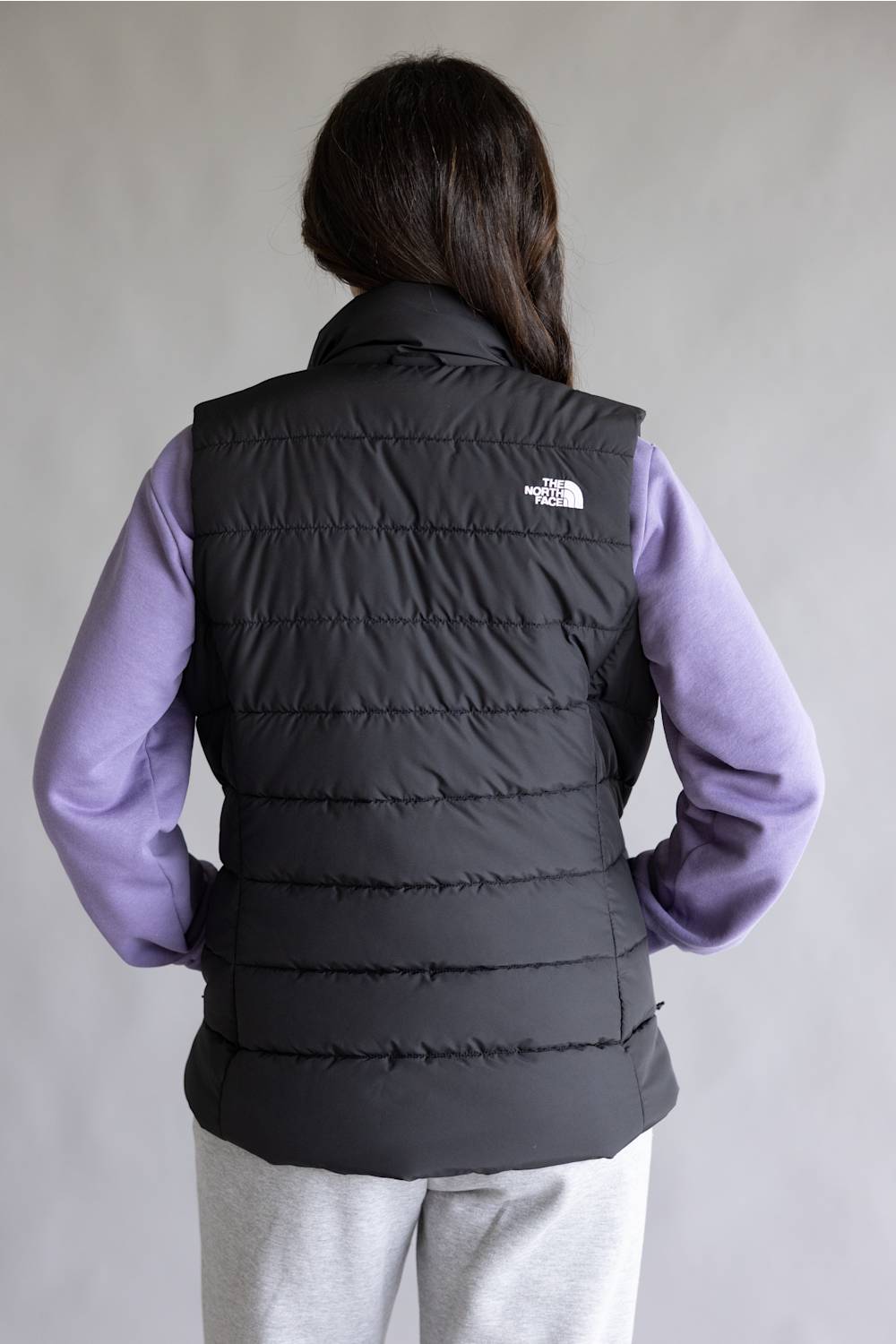 A person shown from the back wearing a black quilted puffer vest over a lavender long-sleeve shirt and light gray sweatpants, against a plain light gray studio background. The The North Face logo is visible on the upper back of the vest.