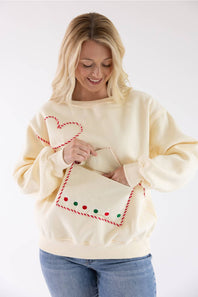 A woman wearing a cream-colored crewneck sweatshirt with a Christmas-themed pocket design, holding a matching cream envelope-shaped purse with red and green accents.