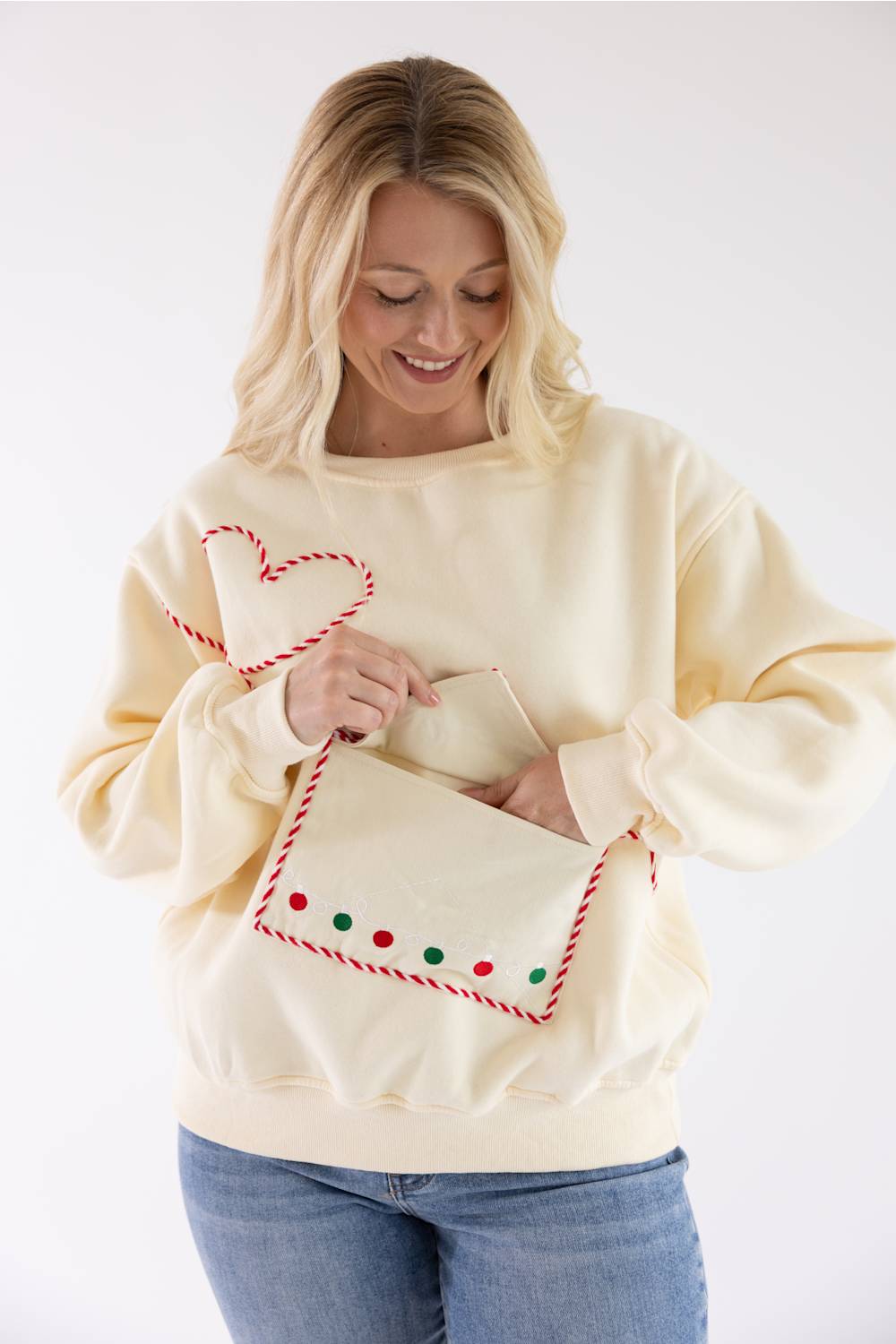 A woman wearing a cream-colored crewneck sweatshirt with a Christmas-themed pocket design, holding a matching cream envelope-shaped purse with red and green accents.