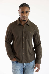 A man wearing a dark brown long-sleeve button-up shirt with a chest pocket and light blue jeans, standing against a plain white background.
