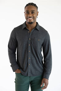 A smiling man wearing a dark charcoal gray long-sleeve button-up shirt with brown buttons and dark green pants, standing against a plain white studio background.