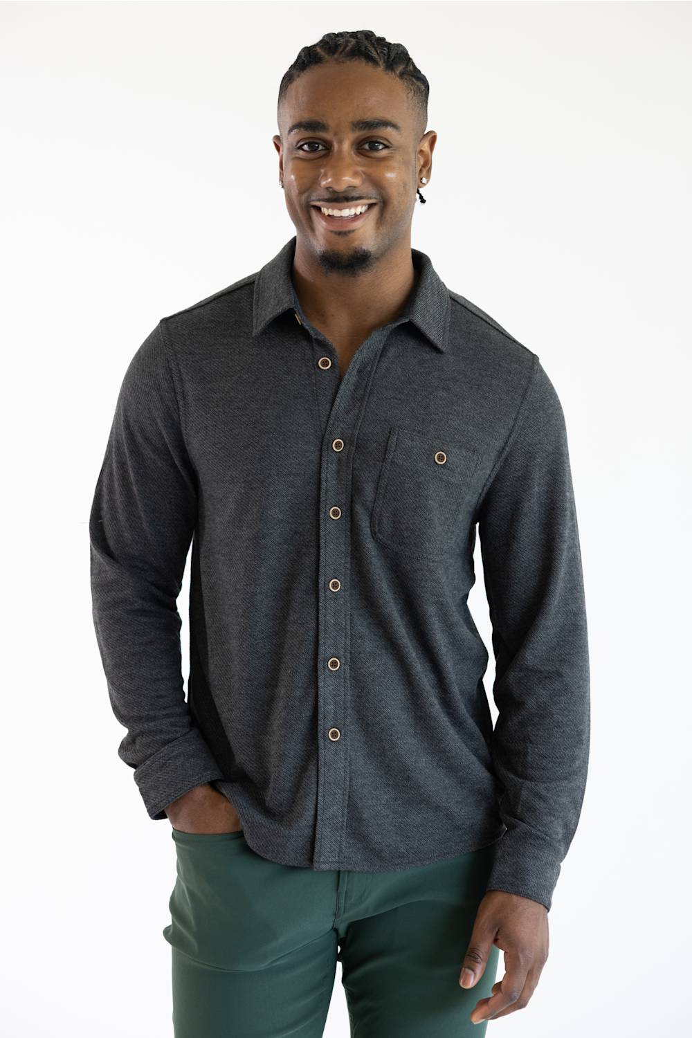 A smiling man wearing a dark charcoal gray long-sleeve button-up shirt with brown buttons and dark green pants, standing against a plain white studio background.