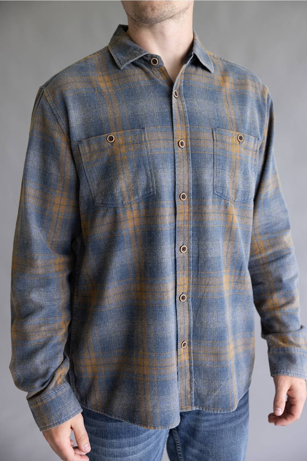 A man wearing a long-sleeve button-up flannel shirt with a blue and brown plaid pattern, paired with blue jeans, shown from the neck down against a neutral gray background.