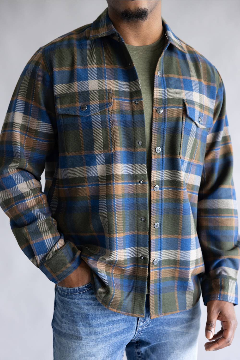 A man wearing a long-sleeve button-up plaid flannel shirt in blue, green, and tan, layered over an olive green t-shirt and paired with blue jeans.