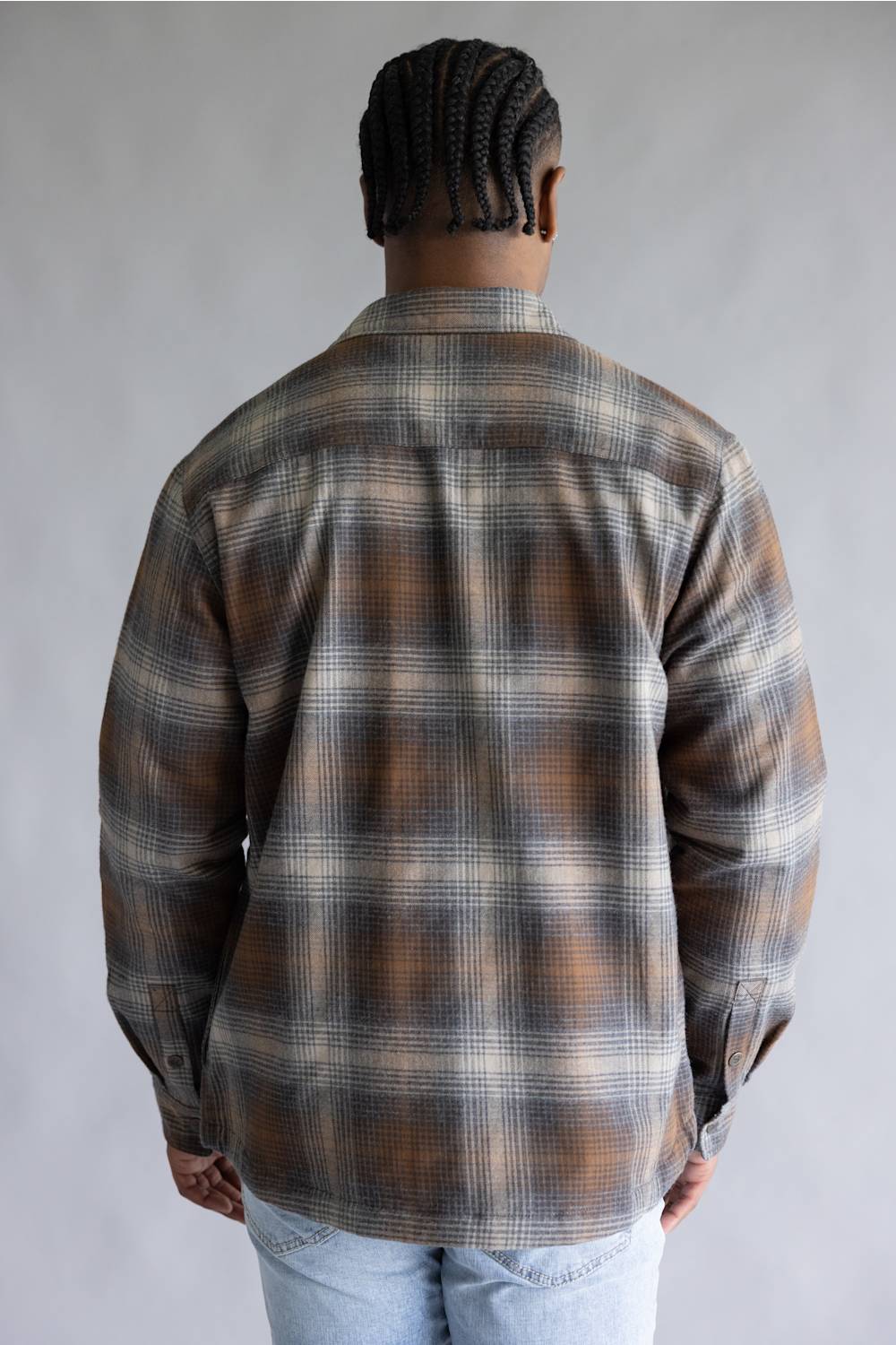 A man viewed from behind wearing a brown, gray, and beige plaid flannel shirt and light blue jeans, standing against a neutral gray studio background.