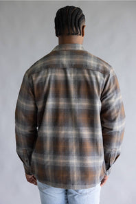 A man viewed from behind wearing a brown, gray, and beige plaid flannel shirt and light blue jeans, standing against a neutral gray studio background.