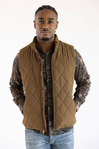 A man wearing a brown diamond-quilted zip-up vest over a camouflage long-sleeve shirt and blue jeans, standing against a plain white background.