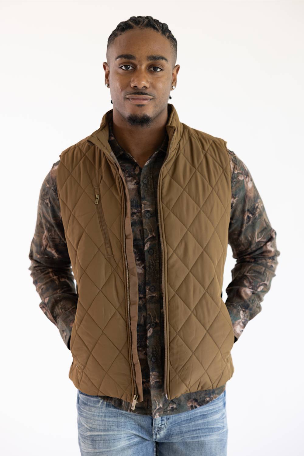 A man wearing a brown diamond-quilted zip-up vest over a camouflage long-sleeve shirt and blue jeans, standing against a plain white background.