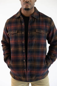 A man wearing a dark brown and black plaid button-up overshirt jacket over a black t-shirt and khaki pants, photographed against a plain white background.
