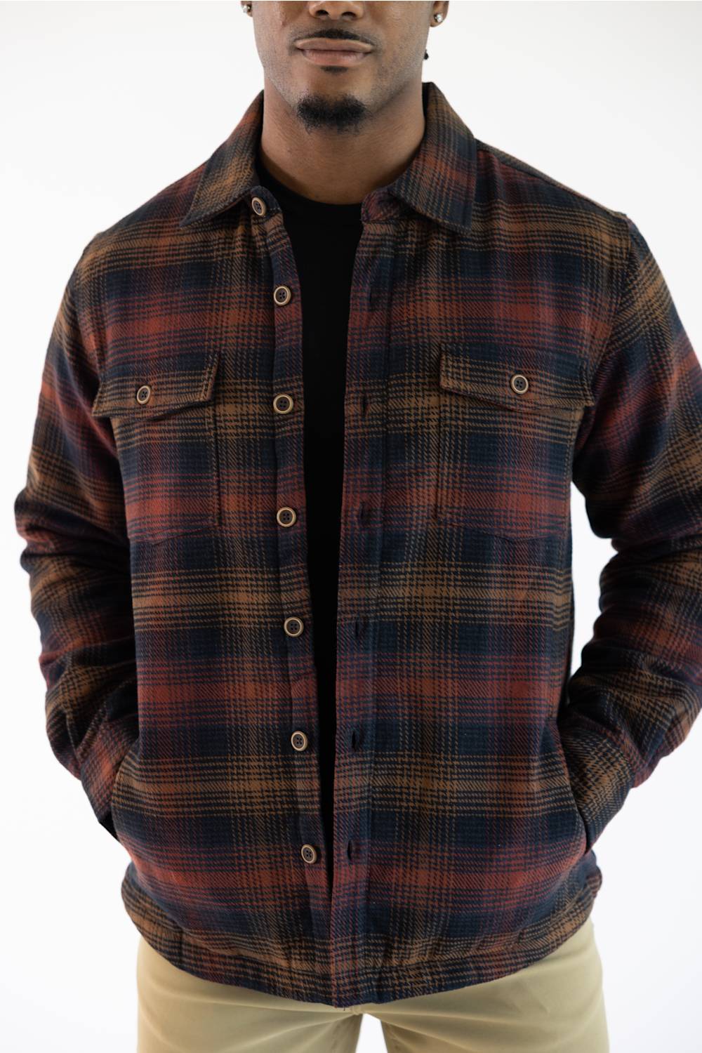 A man wearing a dark brown and black plaid button-up overshirt jacket over a black t-shirt and khaki pants, photographed against a plain white background.