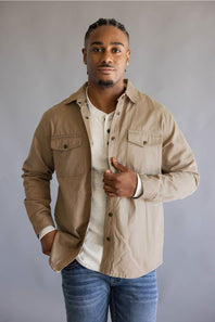 A man wearing a beige button-up shirt jacket over a white henley t-shirt and blue jeans, standing against a neutral gray studio background.