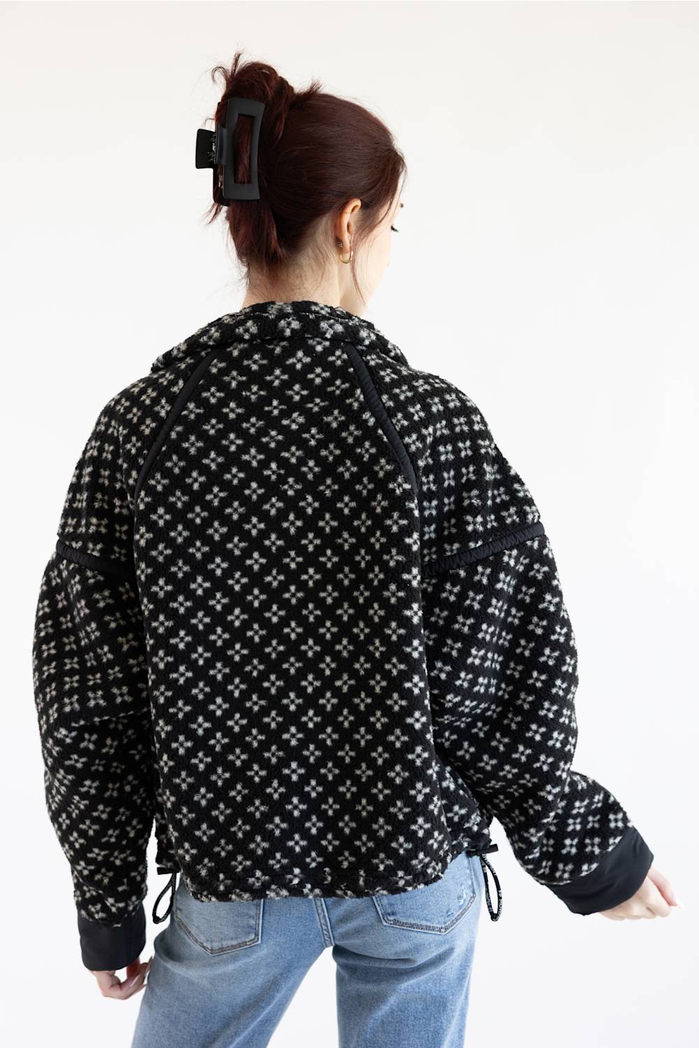 A woman viewed from behind wearing a black and white patterned cropped jacket with drawstrings and light blue jeans, against a plain white background.