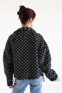 A woman viewed from behind wearing a black and white patterned cropped jacket with drawstrings and light blue jeans, against a plain white background.